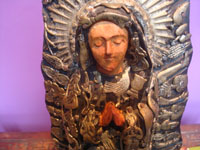 Mexican vintage devotional art, a beautiful woodcarving of Our Lady of Guadalupe decorated with hundreds of milagros, Mexico, c. 1950. Closeup photo of Our Lady's face.