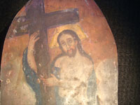 Mexican vintage devotional art, a tin retablo of Christ clutching the cross, c. 1850-1900. The images are hand-painted on tin and are exquisite. The retablo is arch-shaped, resembling a typical stained-glass window in a church. A closeup photo showing Christ clutching his cross.
